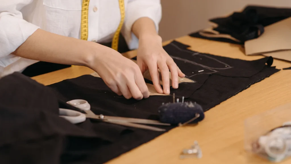 Hands working on marked black fabric beside scissors and a pincushion on a wooden table.