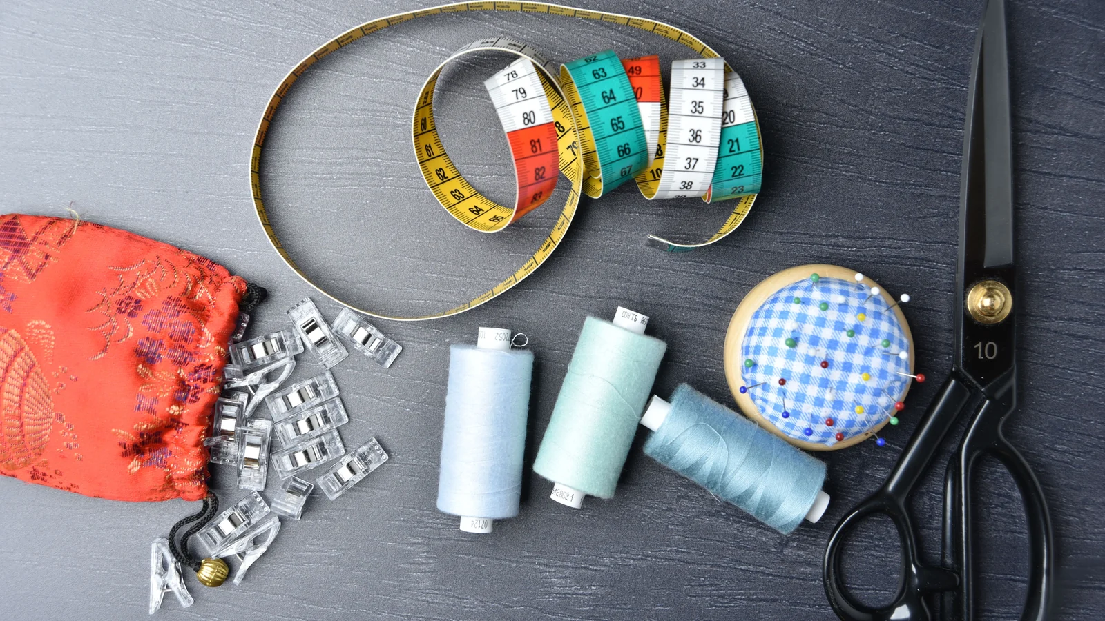Measuring tapes, thread spools, scissors, clips and a round pincushion on a dark surface.